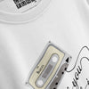 An angled, close-up product shot of a white t-shirt showing a detailed view of a graphic print featuring a vintage cassette tape labeled 'THE BEATLES' with a wire trailing from it towards cursive text, with the brand tag visible near the collar.