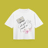 A flat-lay product photograph of a white t-shirt centered on a solid olive-green background. The graphic print features a cassette tape labeled 'The Beatles' connected by a winding wire to the words 'All you need is' and a stylized, flowery 'Love' below, decorated with small illustrations of birds and flowers.