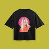 A flat-lay product photograph of a black t-shirt centered on a solid olive-green background. The shirt features a graphic portrait illustration of a blonde woman wearing an orange top, framed by a solid pink circle.