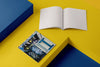 Product photograph of an open, blank sketchbook positioned next to a closed sketchbook featuring a photographic cover design of a graffiti-covered payphone, displayed on a contrasting yellow and blue geometric platform.