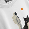 An angled, close-up product shot of a white t-shirt showing a detail of the graphic print, which features an astronaut figure standing beside a black-and-white cat, with a small orange planet in the background and the brand tag visible near the collar.