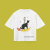 A flat-lay product photograph of a white t-shirt centered on a solid olive-green background. The graphic print features a whimsical scene with a cat standing on a banana that has flames shooting from one end like a rocket, with an astronaut standing on the cat's back. The design includes small stars and planets, the text 'Inspired by a quest for a potassium laden adventure' written below the banana, and the word 'DAYDREAMER' printed at the bottom right.