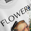 An angled close-up of a white t-shirt graphic, featuring the word 'FLOWERY' in bold black capital letters above a painted depiction of a person having their hair styled with a comb, alongside floral elements and a brand tag visible at the collar.