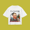 A flat-lay product photograph of a white t-shirt centered on a solid olive-green background. The graphic print features a Vincent van Gogh self-portrait surrounded by colorful flowers, with a hand holding a comb to his hair and another hand near his face, all beneath the word 'FLOWERY' written in bold black text.