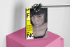Product lifestyle photograph of an art cover sketchbook featuring the GAZNG charcoal portrait with typography, by Nemozine, suspended above a vibrant pink platform against a clean white background.