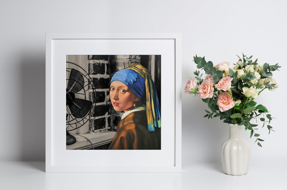  Framed art print of an oil painting of 'girl with a pearl earring' placed next to a vase with flowers on a white surface.
