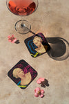 Product lifestyle photograph of two art coasters featuring the IDEAL FEMALE HEAD design by Nemozine, displayed on a textured surface alongside a stemmed glass of pink beverage and scattered pink flower petals.