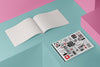 Product photograph of an open, blank sketchbook positioned next to a closed sketchbook featuring a cover adorned with a collage of various stylized illustrations by Nemozine, displayed against a vibrant pink and teal geometric background.