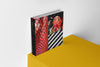 Product photograph of an art sketchbook featuring a triptych graphic design with red tiled textures and stylized characters, displayed on a vibrant yellow platform against a clean white wall.