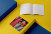 Product photograph of an open, blank sketchbook positioned next to a closed sketchbook featuring a triptych graphic design with red tiled textures and stylized characters, displayed on a contrasting yellow and blue geometric platform.