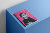 A sketchbook resting on a blue geometric surface, featuring a pink cover with a charcoal illustration of a child in a black shirt surrounded by a repeating pattern of small ants and white circles.