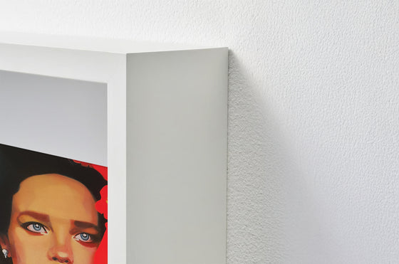 Close-up of a (white shadow box) framed artwork featuring a beautiful womans portrait painting on a textured white wall
