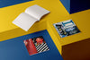Product photograph of an open, blank sketchbook positioned next to two closed sketchbooks with distinct graphic covers, all displayed on a contrasting yellow and blue geometric platform.