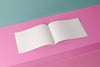 Product lifestyle photograph of a minimalist white notepad with blank pages displayed against a vibrant pink and teal geometric background.