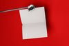 Product lifestyle photograph of a minimalist portrait notepad with blank pages suspended from a metal clamp against a vibrant red background.
