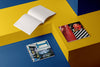 Product photograph of an open, blank sketchbook positioned next to two closed sketchbooks with distinct graphic covers, all displayed on a contrasting yellow and blue geometric platform.