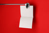 Product lifestyle photograph of a minimalist square notepad with blank pages suspended from a metal clamp against a vibrant red background.
