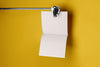 Product lifestyle photograph of a minimalist square notepad with blank pages suspended from a metal clamp against a vibrant yellow background.