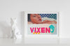 Lifestyle product photograph of the limited edition archival art print VIXEN by Nemozine, displayed in a white frame next to a geometric, white fox figurine on a clean, white surface.