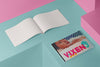 Product photograph of an open, blank sketchbook positioned next to a closed sketchbook featuring a cover adorned  with a provocative still from BLUE IS THE WARMEST COLOUR artwork by Nemozine, displayed against a vibrant pink and teal geometric background.